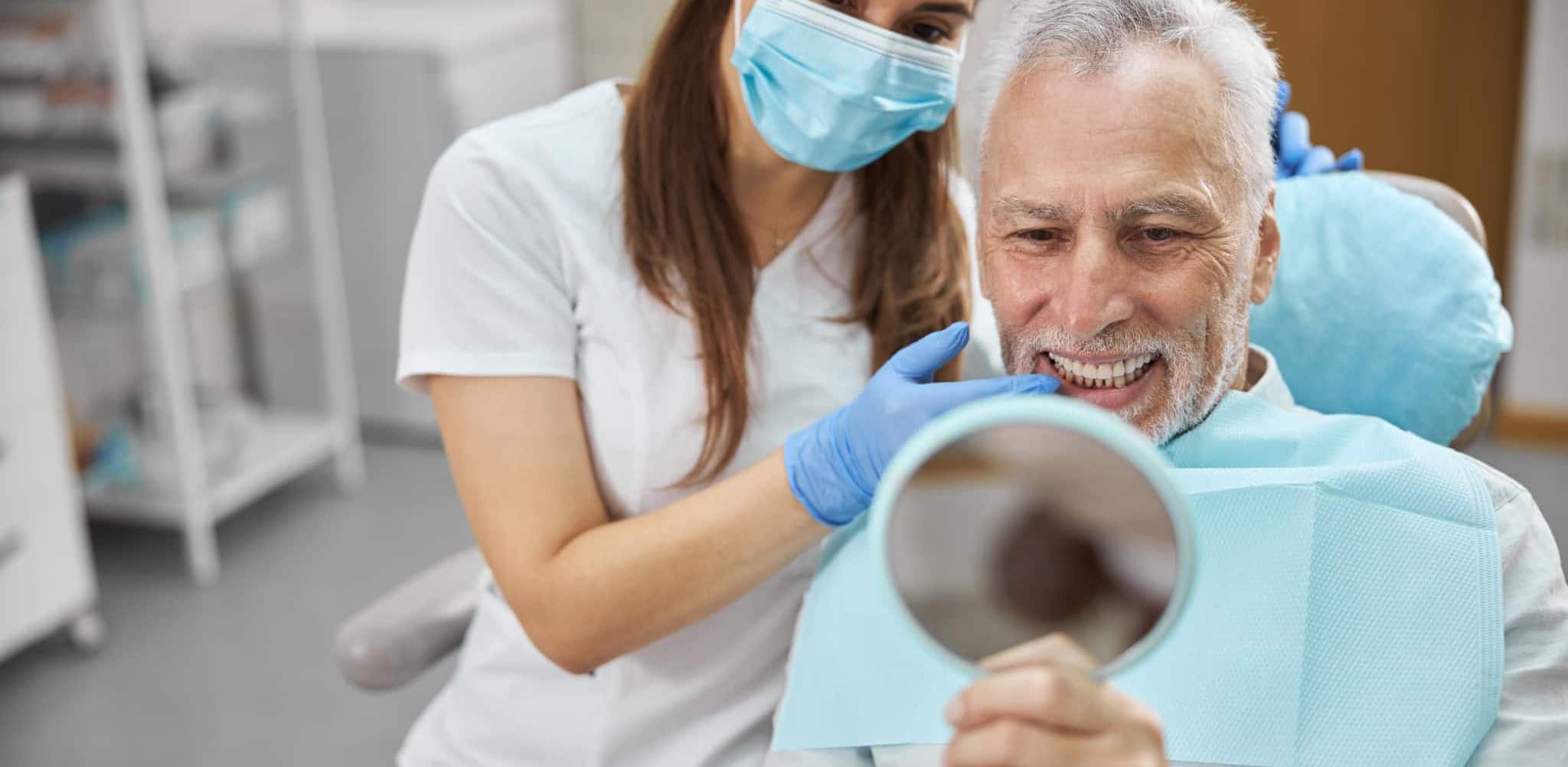 e_r_dental_bg_img_5 A smiling man is checking his new teeth in a mirror with a dentist beside him.